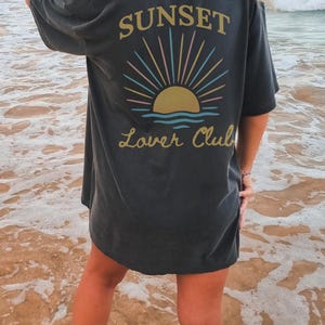 May include: A black oversized t-shirt with a graphic of a sunset and the text "Sunset Lover Club" on the back.