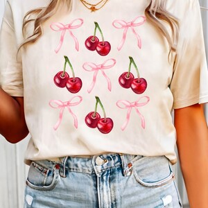 May include: A cream-colored t-shirt with a graphic of pink bows and red cherries.