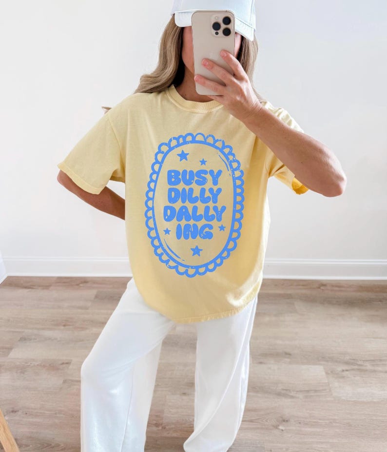 May include: A pale yellow t-shirt featuring the blue text "BUSY DILLY DALLY ING" within a scalloped oval. The shirt is worn with white trousers and a white baseball cap.