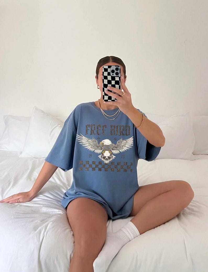 May include: A light blue oversized t-shirt with a graphic print of an eagle with a skull and the text "Free Bird" and "1975".