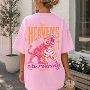 May include: Pink t-shirt with a graphic of a roaring tiger and the text "THE HEAVENS are roaring" in orange. The shirt also features the text "PSALM 19:1".