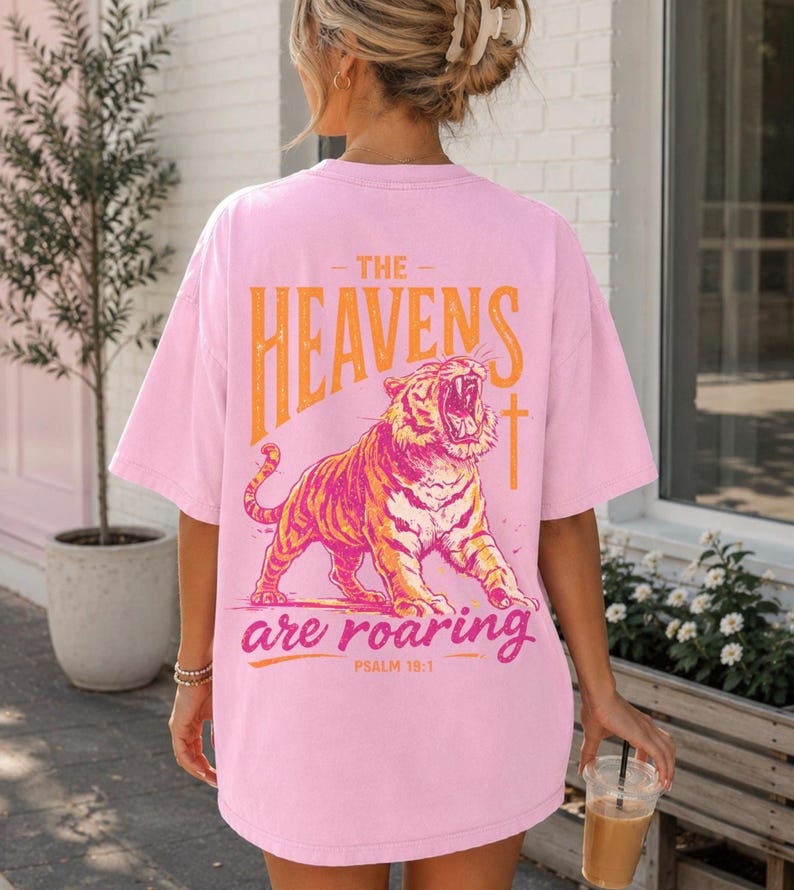 May include: Pink t-shirt with a graphic of a roaring tiger and the text "THE HEAVENS are roaring" in orange. The shirt also features the text "PSALM 19:1".