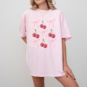 May include: A pink oversized t-shirt with a pattern of cherries with pink bows.
