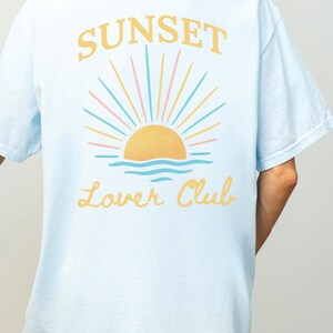 May include: Light blue t-shirt with a graphic of a sunset over the ocean and the text "Sunset Lower Club" in yellow.