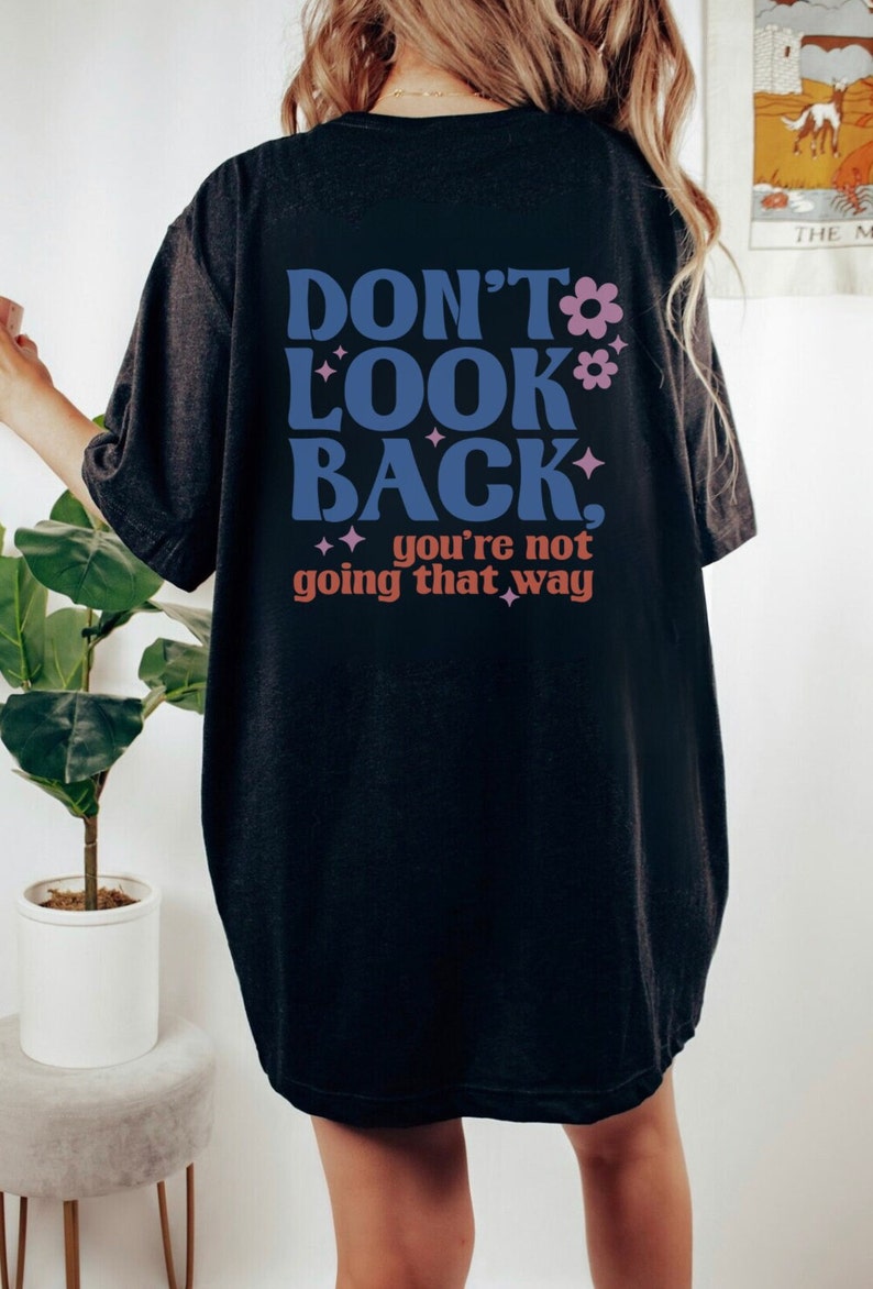 May include: Black oversized t-shirt with the text "Don't Look Back, you're not going that way" in blue, pink, and orange lettering with pink flowers and glitter accents.