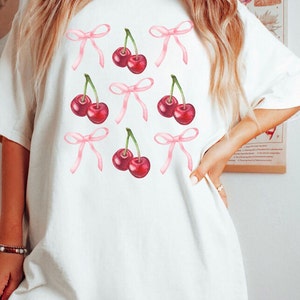 May include: White t-shirt featuring a pattern of red cherries and pink bows. The cherries have green stems. The bows are a light pink color. The shirt is a casual, oversized style.