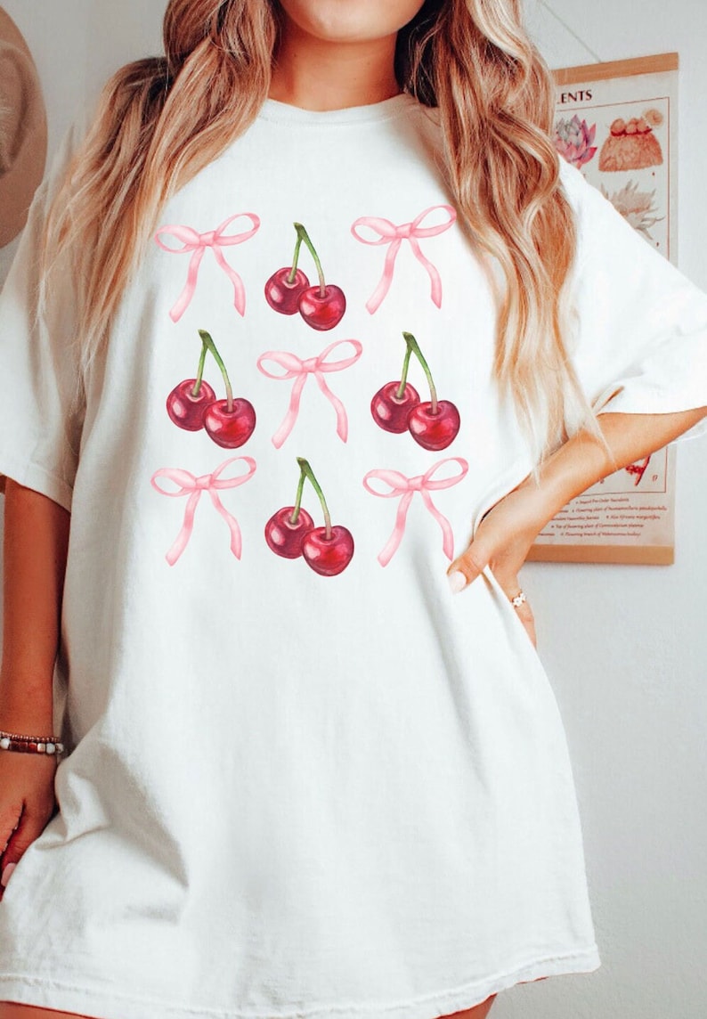 May include: White t-shirt featuring a pattern of red cherries and pink bows. The cherries have green stems. The bows are a light pink color. The shirt is a casual, oversized style.