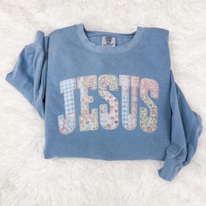 May include: A light blue sweatshirt with the word "JESUS" appliquéd across the front. The letters are made of patchwork fabric with floral and gingham patterns. The sweatshirt has a crew neck and long sleeves.
