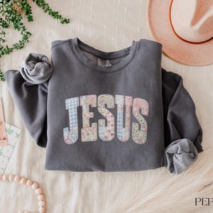 May include: A charcoal gray sweatshirt with the word "JESUS" in a patchwork design of floral and gingham patterns. The long-sleeved top is folded on a cream-colored surface, with a hat and greenery in the background.