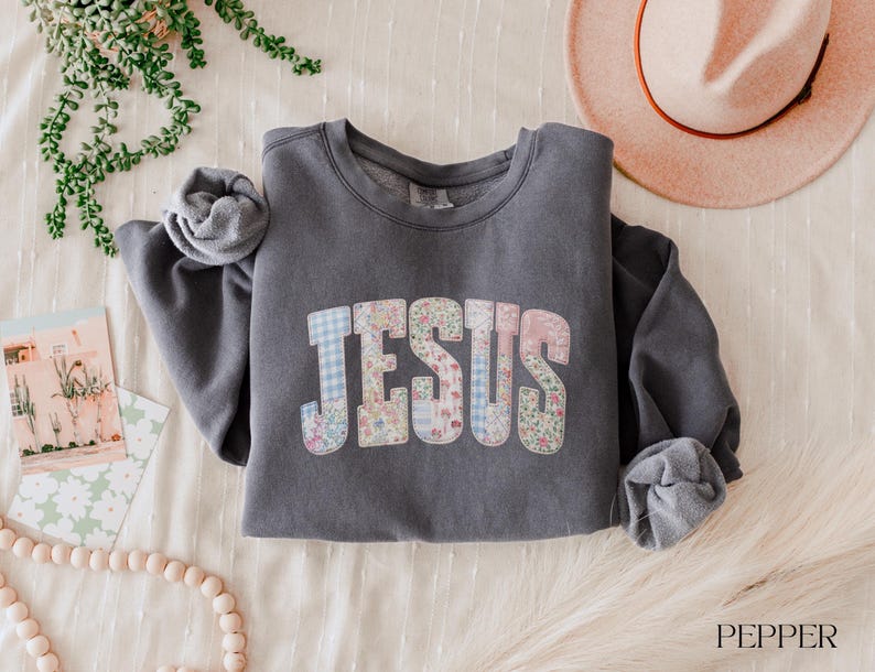 May include: A charcoal gray sweatshirt with the word "JESUS" in a patchwork design of floral and gingham patterns. The long-sleeved top is folded on a cream-colored surface, with a hat and greenery in the background.