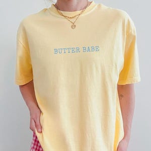 May include: A light yellow t-shirt with the text "BUTTER BABE" in light blue. The shirt has short sleeves and a crew neck. A gold necklace with a heart pendant is visible.