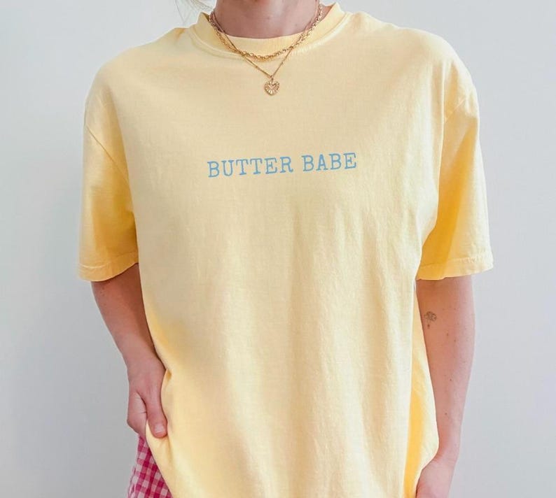 May include: A light yellow t-shirt with the text "BUTTER BABE" in light blue. The shirt has short sleeves and a crew neck. A gold necklace with a heart pendant is visible.