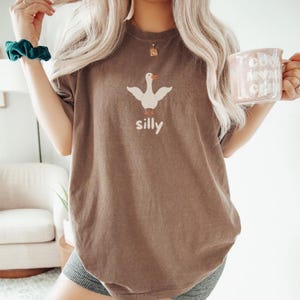 May include: A brown oversized t-shirt with a white goose graphic and the word "silly" printed below it. The person is wearing grey shorts and holding a white mug with the text "cool mom club" printed on it.