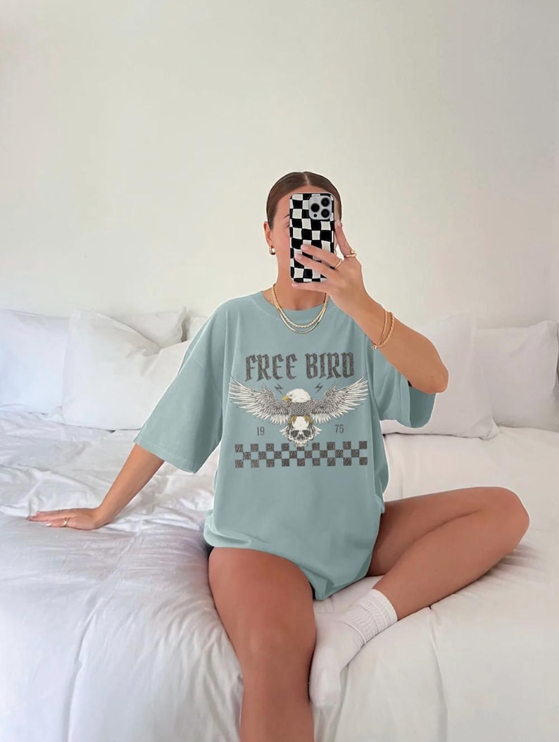 May include: A light blue t-shirt with a graphic of an eagle with a skull and crossbones. The text "FREE BIRD" is printed above the graphic. The graphic is surrounded by a checkered pattern.