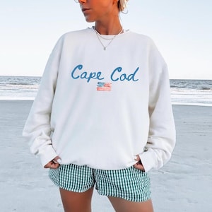 Op de afbeelding: Crèmekleurige sweatshirt met de woorden "Cape Cod" in blauw schrift en een kleine Amerikaanse vlag. De sweatshirt wordt gedragen met groen-wit gestreepte shorts op een strand.