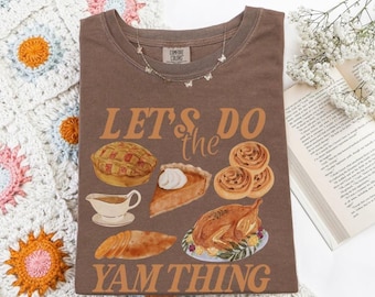 May include: A brown Comfort Colors t-shirt with the phrase "LET'S DO the YAM THING" in orange lettering. The shirt features illustrations of Thanksgiving foods like pie, turkey, and rolls. A delicate butterfly necklace rests on the shirt.