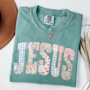 May include: A teal Comfort Colors t-shirt with the word "JESUS" appliqued in floral and gingham fabric. A silver necklace with a small pendant rests on the shirt. The shirt is folded, with a brown hat and dried palm leaf in the background.