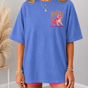 May include: Flo Blue short-sleeved t-shirt with a tiger graphic. The graphic includes the text "THE HEAVENS are roaring". The shirt is a solid blue color. The text "FLO BLUE" is printed at the bottom of the shirt.