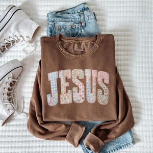 May include: A brown sweatshirt with the word "JESUS" in a patchwork design of floral and gingham patterns. The sweatshirt is paired with blue jeans, a gold chain necklace, and white high-top sneakers.