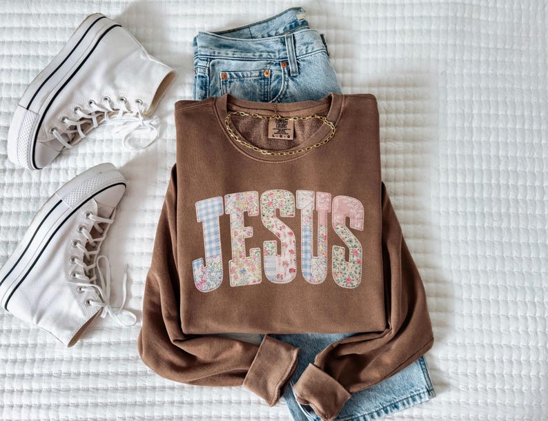 May include: A brown sweatshirt with the word "JESUS" in a patchwork design of floral and gingham patterns. The sweatshirt is paired with blue jeans, a gold chain necklace, and white high-top sneakers.