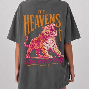May include: Dark gray t-shirt featuring a roaring tiger illustration in orange and pink. The text "THE HEAVENS are ROARING" and "PSALM 19:1" are printed above and below the tiger design.