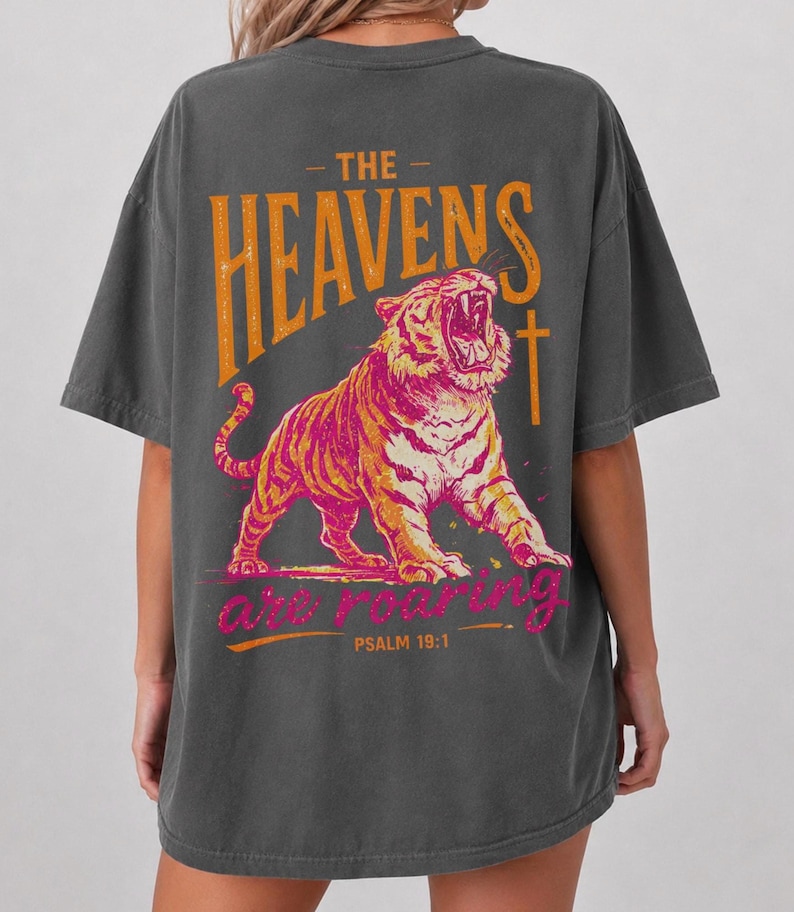 May include: Dark gray t-shirt featuring a roaring tiger illustration in orange and pink. The text "THE HEAVENS are ROARING" and "PSALM 19:1" are printed above and below the tiger design.