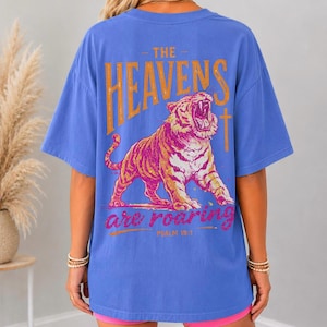 May include: Blue t-shirt with a distressed orange and pink graphic of a roaring tiger and the text "THE HEAVENS are roaring" and "PSALM 19:1". The shirt has short sleeves and a relaxed fit.