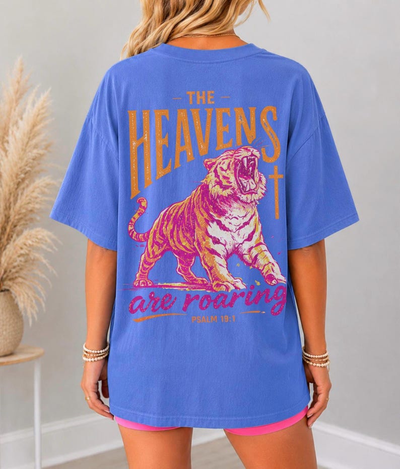 May include: Blue t-shirt with a distressed orange and pink graphic of a roaring tiger and the text "THE HEAVENS are roaring" and "PSALM 19:1". The shirt has short sleeves and a relaxed fit.