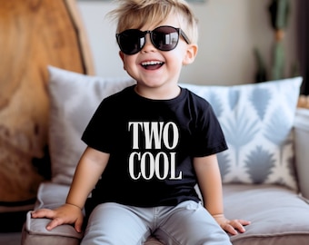 May include: A young child wearing a black t-shirt with the text "TWO COOL" in white letters. The child is wearing sunglasses and smiling.