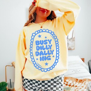 May include: A pale yellow sweatshirt featuring the text "BUSY DILLY DALLY ING" in a blue oval design. The person is wearing black and white checkered shorts and a green cap. The sweatshirt has long sleeves.