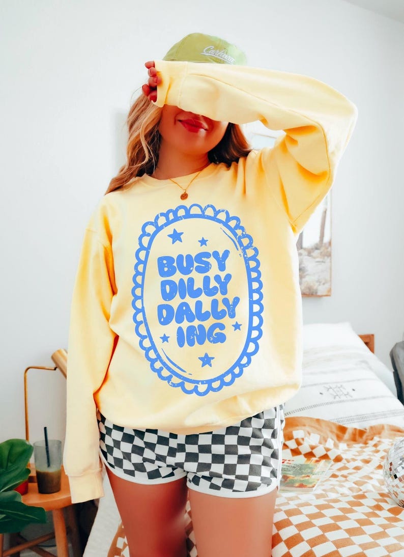 May include: A pale yellow sweatshirt featuring the text "BUSY DILLY DALLY ING" in a blue oval design. The person is wearing black and white checkered shorts and a green cap. The sweatshirt has long sleeves.
