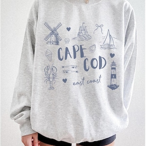 Puede incluir: Sudadera gris claro con un diseño gráfico azul que presenta un molino de viento, un castillo, un velero, un helado, una langosta, peces, un faro y las palabras "CAPE COD east coast".