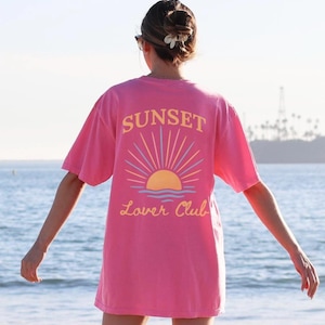 以下が含まれることがあります： 夕焼けと「Sunset Lover Club」の文字がプリントされたピンクのTシャツです。