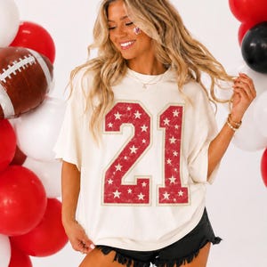 May include: An off-white short-sleeve t-shirt featuring the number 21 in a large, distressed red font filled with small white stars. The shirt has a relaxed fit and is paired with black denim shorts that have a frayed, raw-edge hemline.
