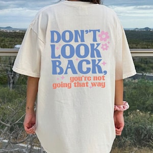 May include: Ivory colored t-shirt with a blue and pink graphic that says "Don't Look Back, you're not going that way".