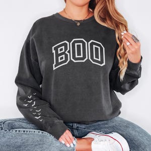 May include: Dark gray sweatshirt with the word "BOO" in white block letters across the chest. The left sleeve features a pattern of white bats. The sweatshirt is a casual, comfortable style.