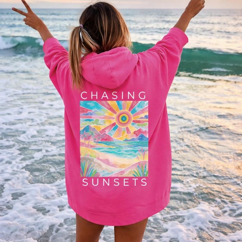 Op de afbeelding: Een felroze hoodie met een aquarel-afbeelding op de rug. De afbeelding toont een kleurrijke zonsondergang met de woorden "CHASING SUNSETS" in het wit. De hoodie heeft een capuchon en lange mouwen.