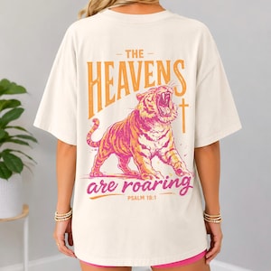 May include: Cream-colored t-shirt featuring a roaring tiger graphic in shades of orange and pink. The text "THE HEAVENS are roaring" is above the tiger, with "PSALM 19:1" below. The shirt has short sleeves and a relaxed fit.