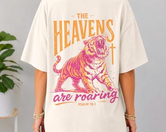 May include: Cream-colored t-shirt featuring a roaring tiger graphic in shades of orange and pink. The text "THE HEAVENS are roaring" is above the tiger, with "PSALM 19:1" below. The shirt has short sleeves and a relaxed fit.