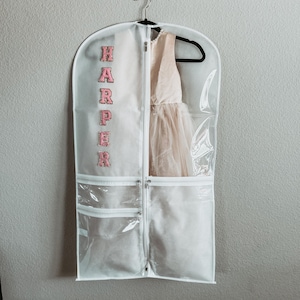 May include: A white garment bag with a clear front panel, hanging on a black hanger. The bag has the name "HARPER" in pink, floral-patterned letters. Inside, a light pink dress is visible.