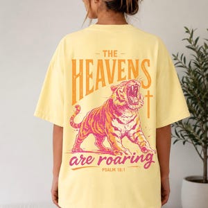 May include: Yellow t-shirt with a graphic of a roaring tiger in pink and orange. The text "THE HEAVENS are ROARING" is above and below the tiger, with "PSALM 19:1" below the text. The shirt is a light yellow color.