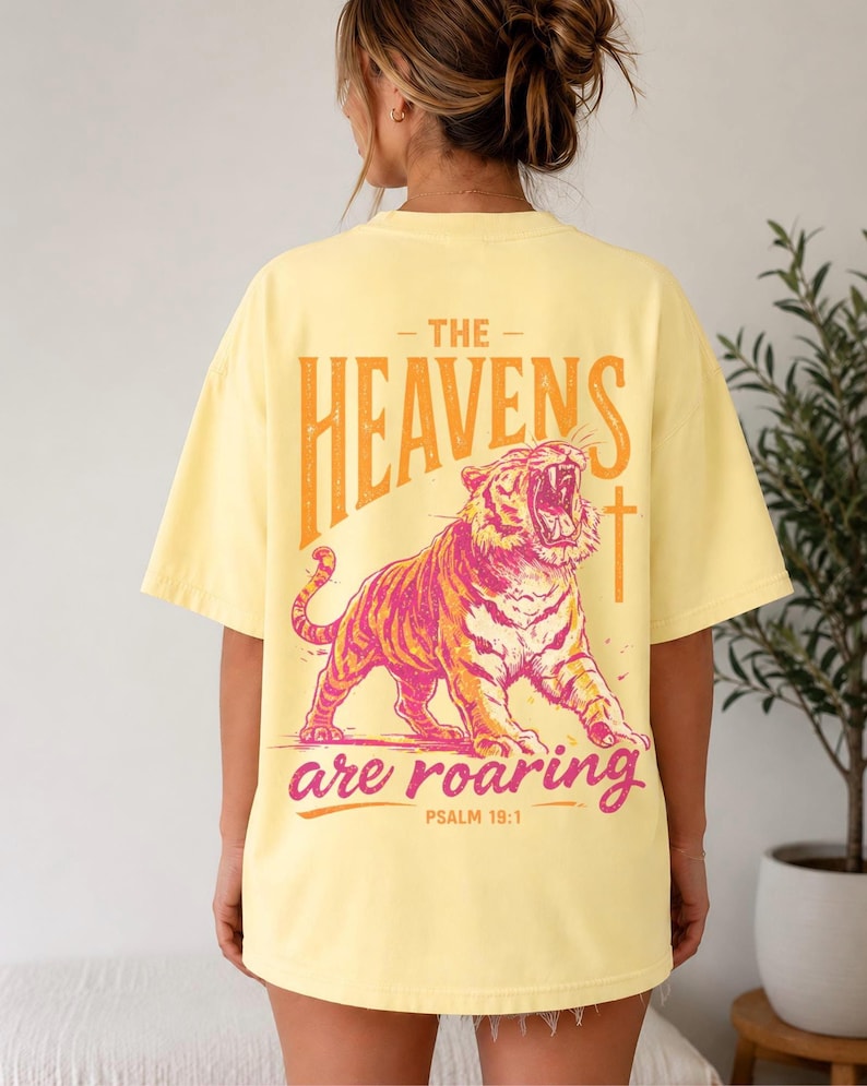 May include: Yellow t-shirt with a graphic of a roaring tiger in pink and orange. The text "THE HEAVENS are ROARING" is above and below the tiger, with "PSALM 19:1" below the text. The shirt is a light yellow color.