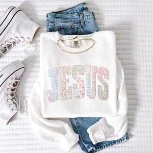 May include: A white sweatshirt with the word "JESUS" in pastel floral and plaid letters. The sweatshirt is paired with blue jeans, a gold chain, and white high-top sneakers. The items are arranged on a white, textured surface.