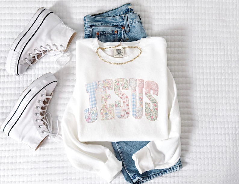 May include: A white sweatshirt with the word "JESUS" in pastel floral and plaid letters. The sweatshirt is paired with blue jeans, a gold chain, and white high-top sneakers. The items are arranged on a white, textured surface.