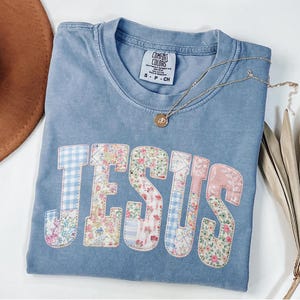 Könnte beinhalten: Hellblaues Comfort Colors T-Shirt mit dem Wort "JESUS" in einem Patchwork-Design auf der Vorderseite appliziert. Die Buchstaben weisen Blumen- und Vichy-Muster auf. Eine goldene Halskette mit Anhänger liegt auf dem Shirt.