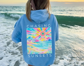 May include: A light blue hoodie with a colorful sunset graphic on the back. The graphic features a sun, mountains, and ocean with the words "CHASING SUNSETS". The hoodie is made of a soft material.