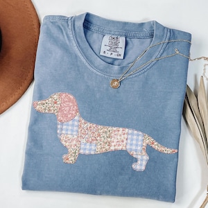 May include: A light blue Comfort Colors t-shirt featuring a patchwork dachshund design. The dachshund is made of floral and gingham fabrics in pink, blue, and white. A gold necklace with a pendant rests on the shirt. The shirt has the text "S-P-CH".