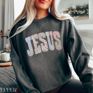 May include: Dark gray sweatshirt with the word "JESUS" in a patchwork design of floral and gingham patterns. The sweatshirt is a crew neck style and has long sleeves. The word "PEPPER" is printed at the bottom left.