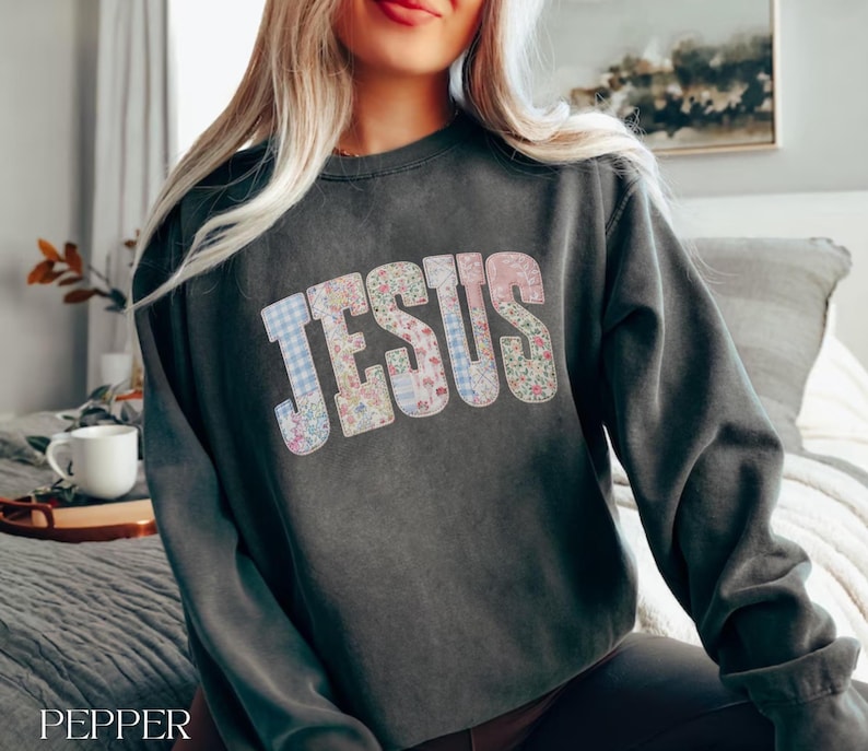 May include: Dark gray sweatshirt with the word "JESUS" in a patchwork design of floral and gingham patterns. The sweatshirt is a crew neck style and has long sleeves. The word "PEPPER" is printed at the bottom left.