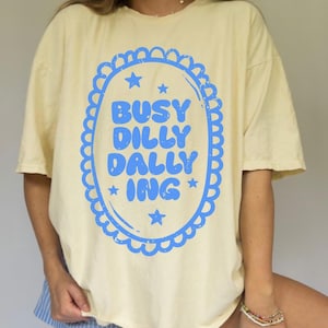 May include: A pale yellow t-shirt featuring a blue oval design with the words "BUSY DILLY DALLY ING". The design is framed by a scalloped border and stars. The shirt has short sleeves.